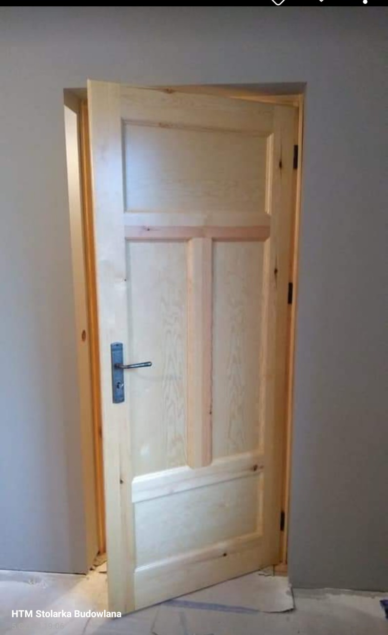 Surowe drewniane drzwi wewnętrzne, trzyskrzydłowe, z prostym metalowym uchwytem, częściowo otwarte, widoczne zawiasy i ościeżnica, na podłodze karton zabezpieczający.