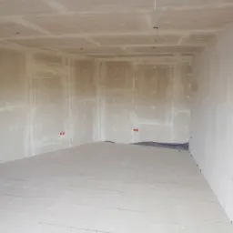 Surowe wnętrze pokoju z płytami kartonowo-gipsowymi na ścianach i suficie, podłoga zabezpieczona folią, widoczne otwory na gniazdka elektryczne.