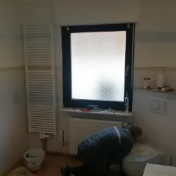 Hydraulik w trakcie montażu lub naprawy spłuczki w łazience z białymi płytkami, widoczny grzejnik, okno z matową szybą i ślady po malowaniu na ścianie.