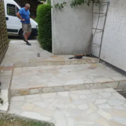 Ułożona ścieżka z nieregularnych kamieni piaskowca prowadząca do budynku, z widocznym pracownikiem w tle i narzędziami na schodach.