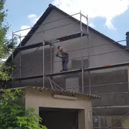 Pracownik na rusztowaniu czyści elewację domu przed ociepleniem przy użyciu myjki ciśnieniowej, widoczny fragment elewacji po czyszczeniu.