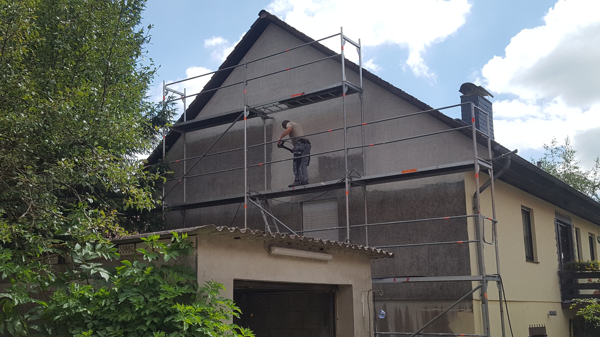 Pracownik na rusztowaniu czyści elewację domu przed ociepleniem przy użyciu myjki ciśnieniowej, widoczny fragment elewacji po czyszczeniu.