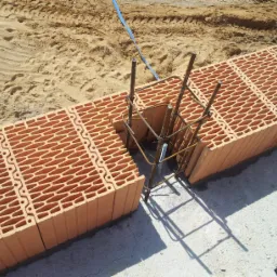Budowa ściany z bloczków ceramicznych na fundamencie, zbrojenie w narożniku, piasek w tle.