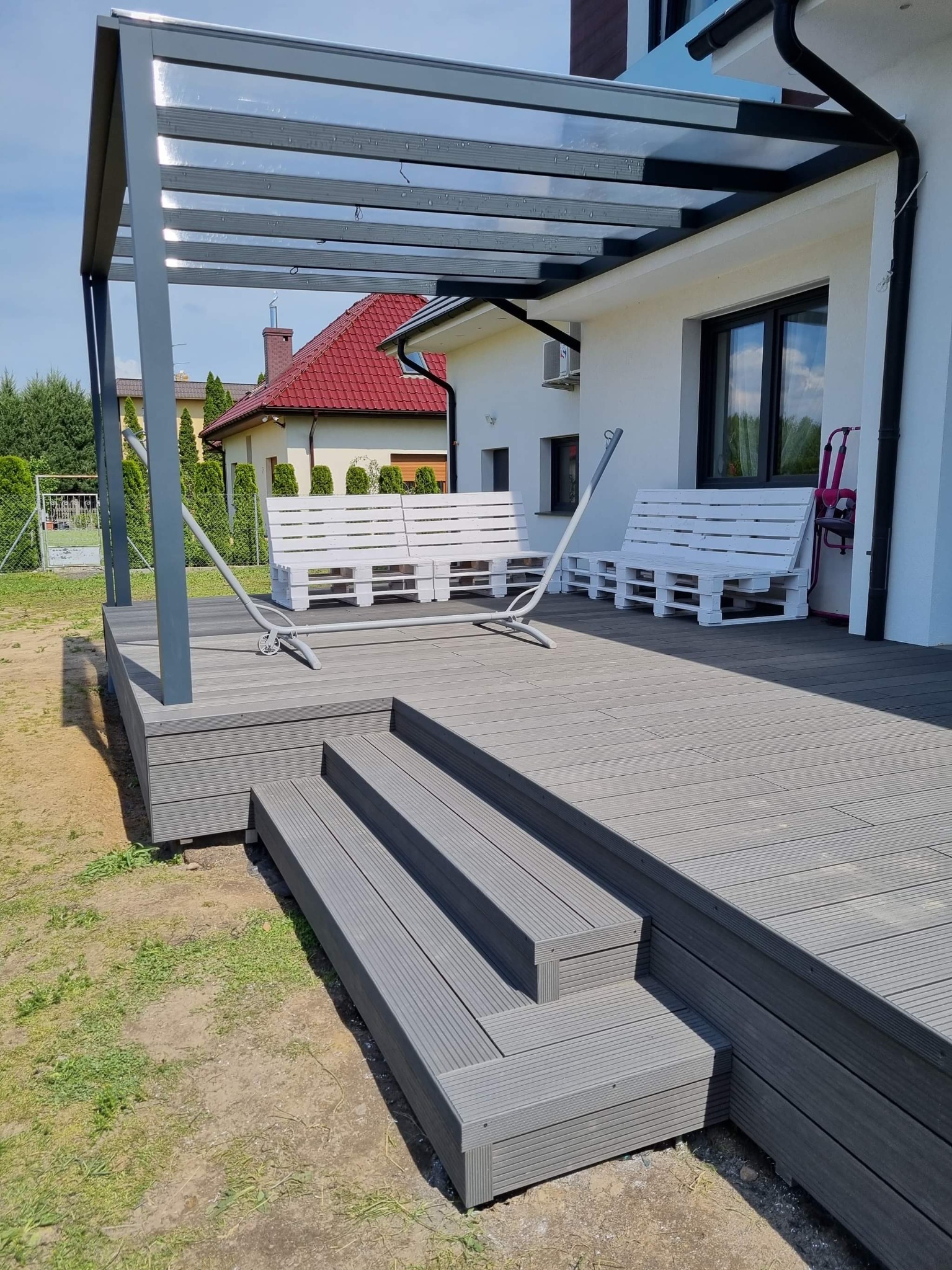 Nowoczesny taras z desek kompozytowych w odcieniach szarości, zadaszony metalową pergolą z przezroczystym poliwęglanem, prowadzący do domu z białymi ławeczkami z palet.