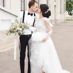 Para młoda w pocałunku na tle jasnego budynku; panna młoda w białej sukni z długim rękawem i kwiatowym wzorem, pan młody w czarnych spodniach, białej koszuli z muchą i szelkami, trzyma bukiet kwiatów.