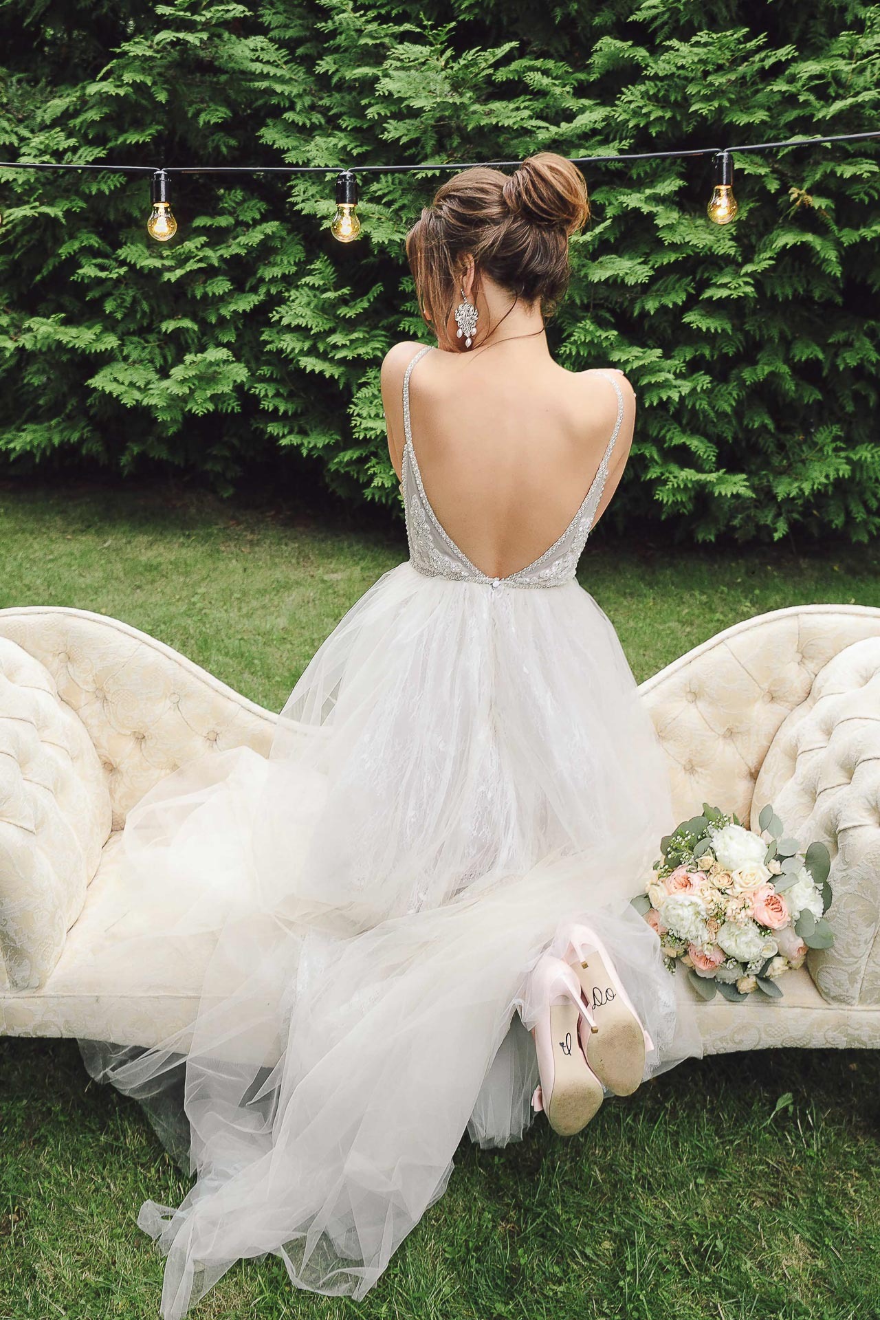 Panna młoda w sukni ślubnej z odkrytymi plecami siedzi na kremowej sofie na tle zieleni, obok leżą buty ślubne z napisem i bukiet kwiatów.