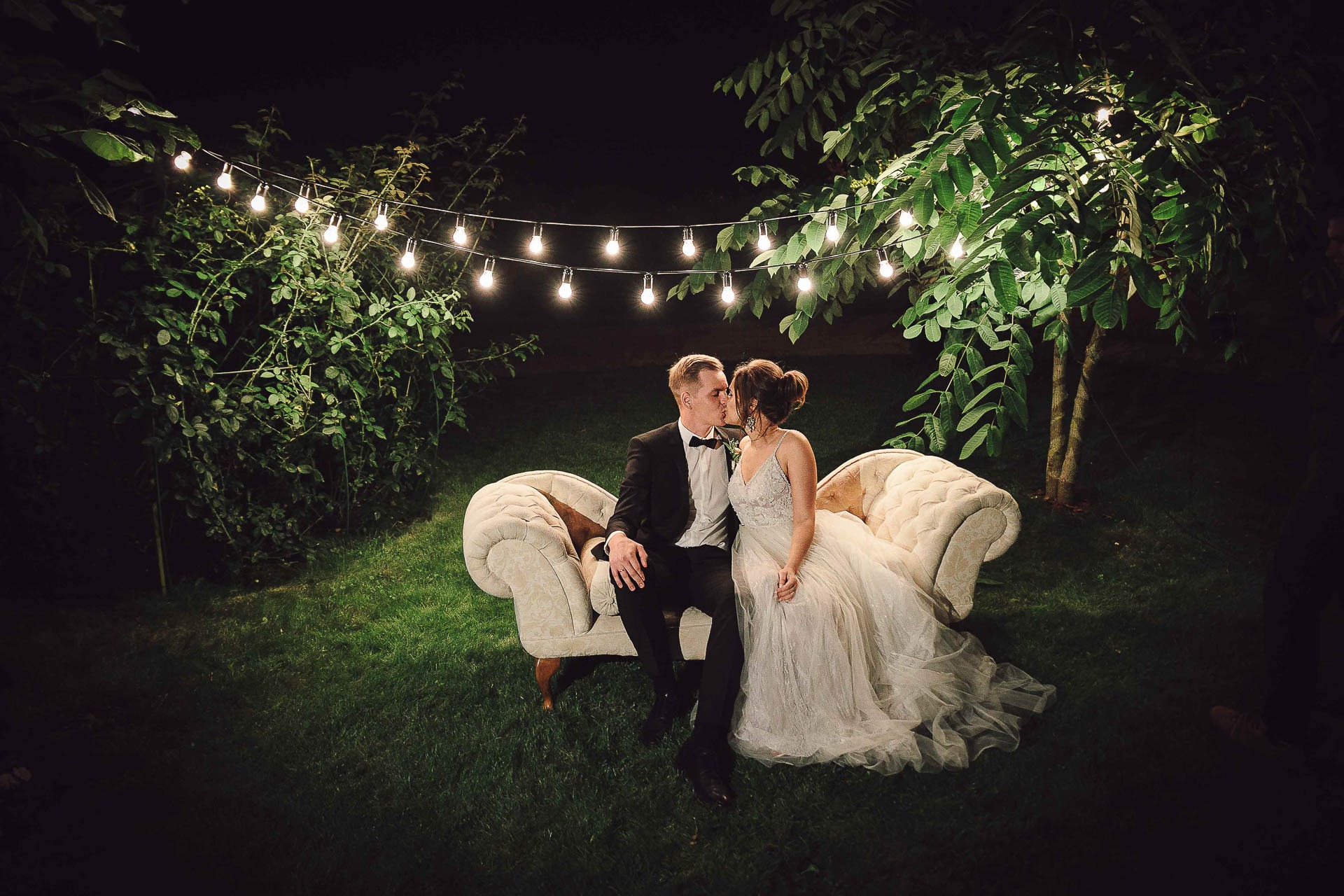 Para młoda w pocałunku siedzi na kremowej sofie na tle drzew i girlandy światełek, on w czarnym smokingu z muszką, ona w białej sukni z koronkową górą.