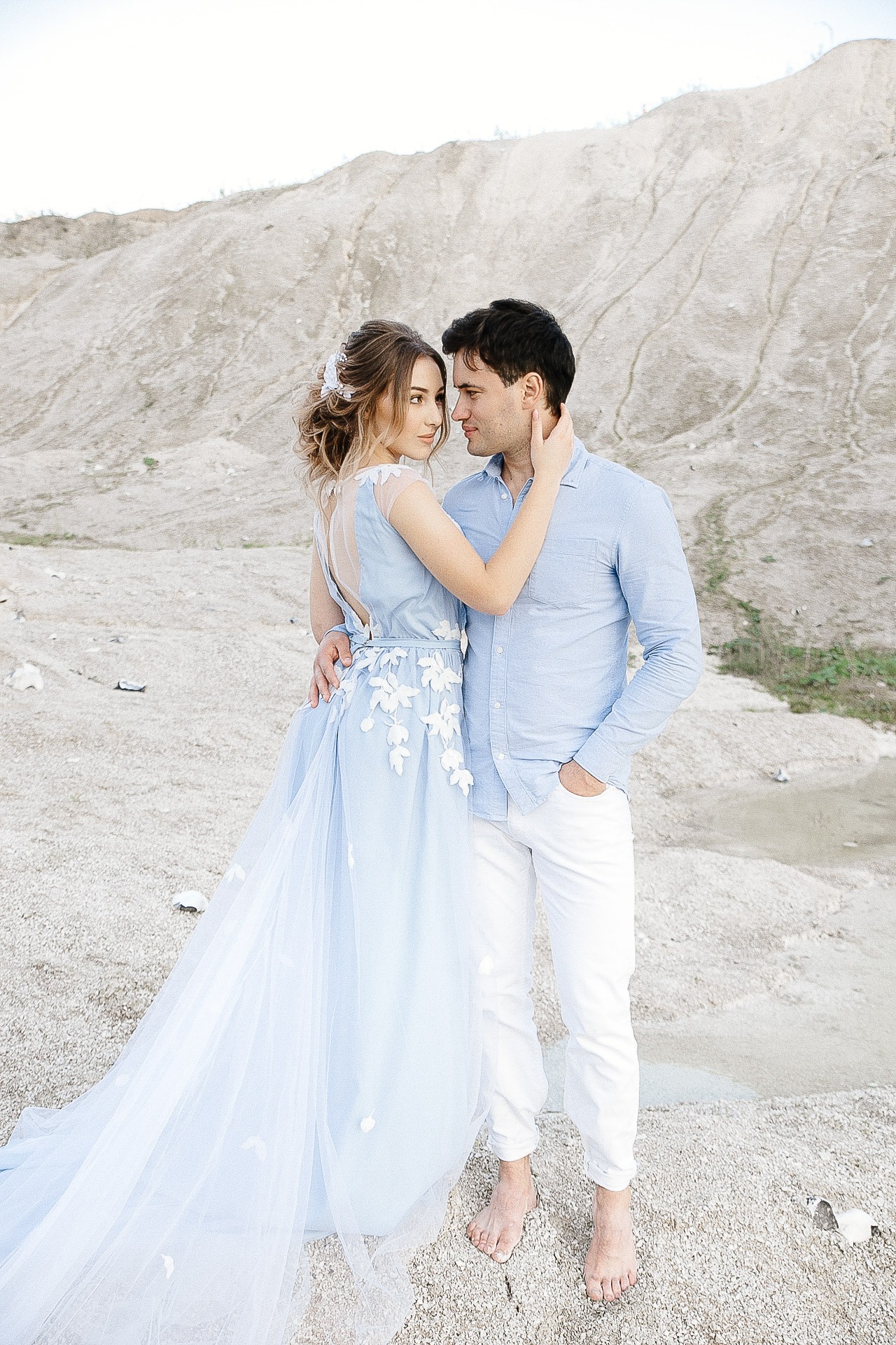 Para narzeczeńska w błękitnych strojach pozuje na tle piaszczystego wzgórza; kobieta w zwiewnej sukni z aplikacjami kwiatowymi, mężczyzna w lnianej koszuli i białych spodni, oboje boso.