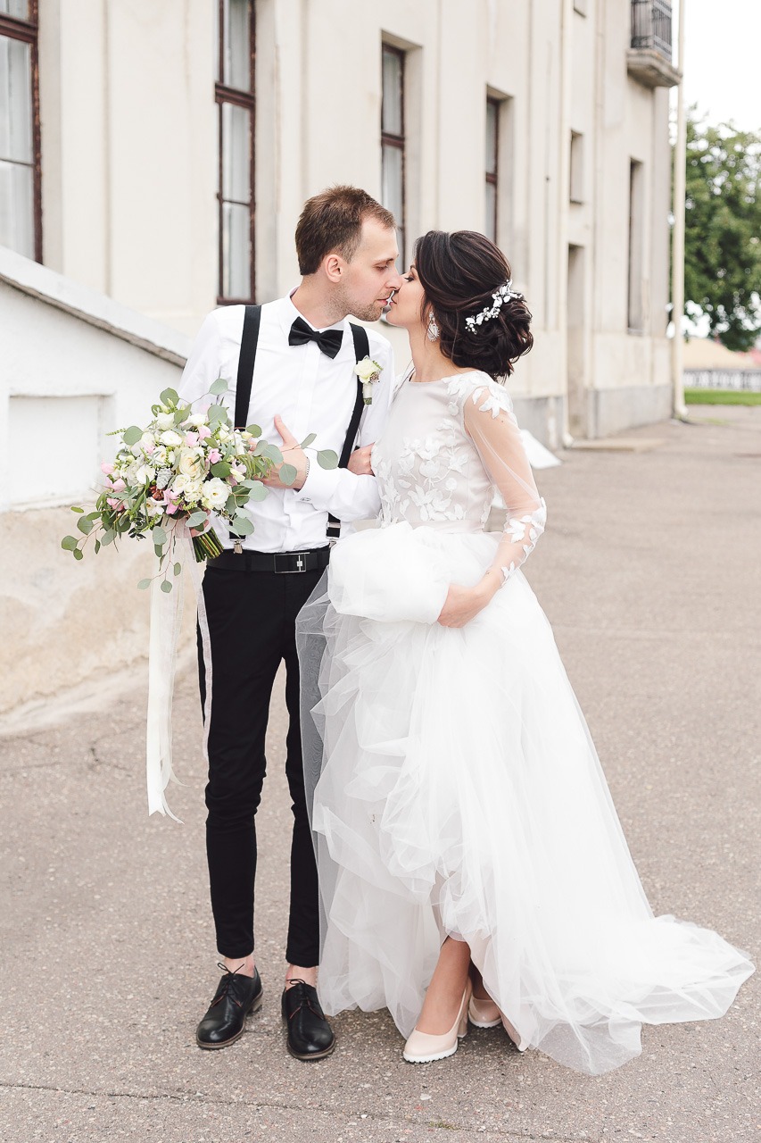 Para młoda w pocałunku na tle jasnego budynku; panna młoda w białej sukni z długim rękawem i kwiatowym wzorem, pan młody w czarnych spodniach, białej koszuli z muchą i szelkami, trzyma bukiet kwiatów.