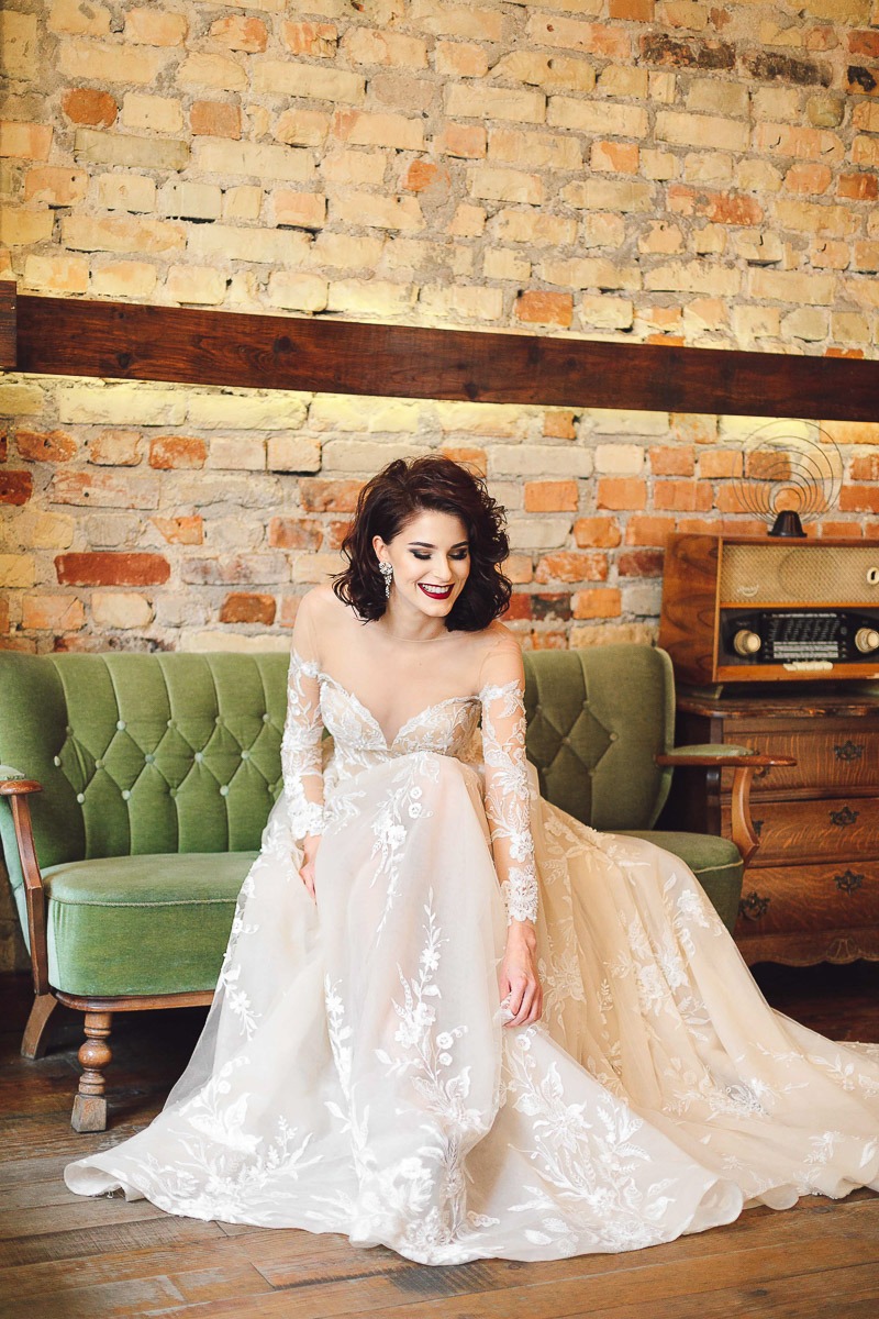 Uśmiechnięta panna młoda w koronkowej sukni siedzi na zielonej sofie na tle ceglanej ściany i starego radia.