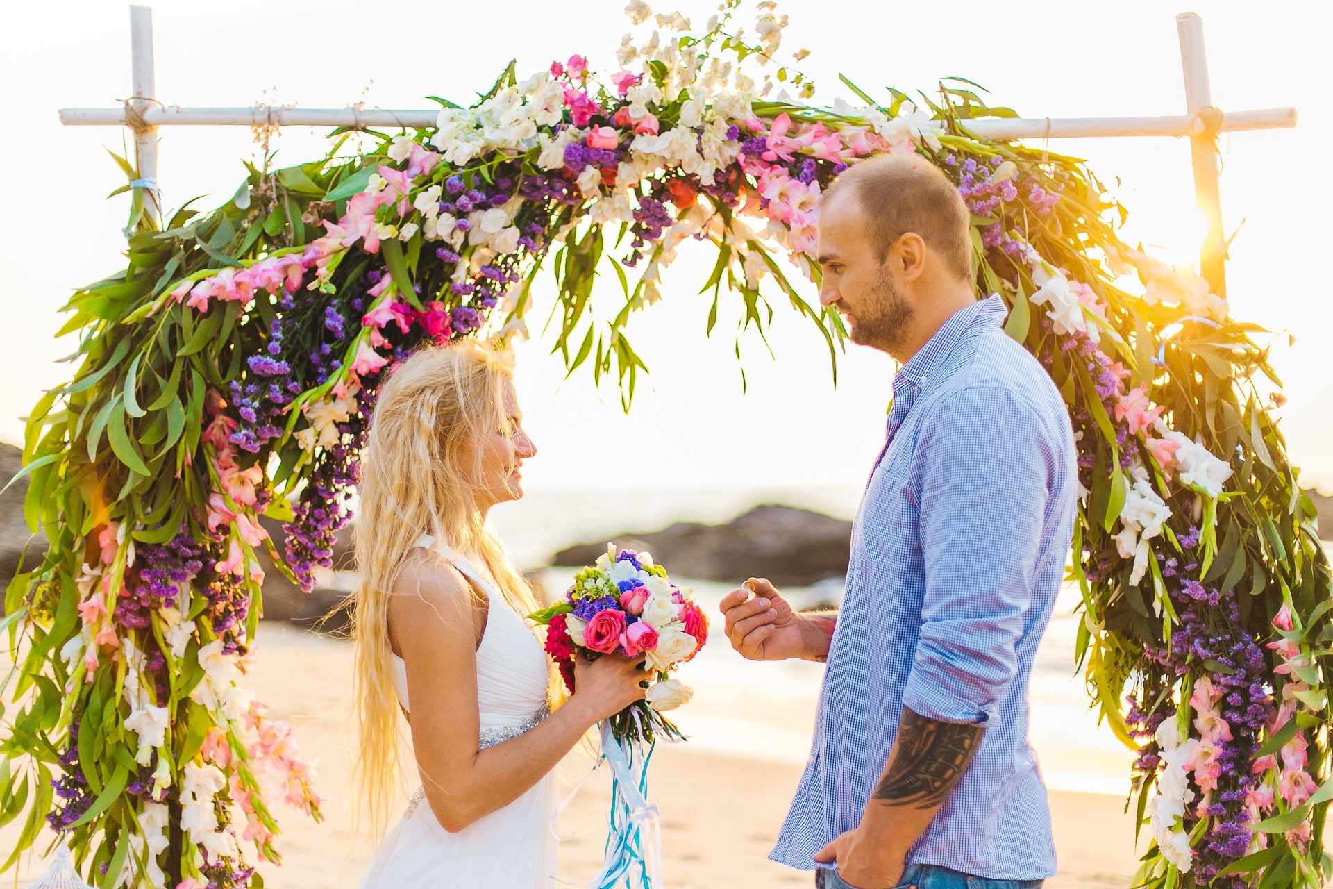 Para na plaży podczas ceremonii ślubnej pod łukiem z kwiatów, mężczyzna wręcza pierścionek kobiecie.