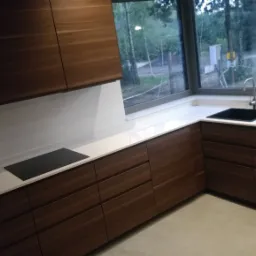 Nowoczesna kuchnia z białym, narożnym blatem i drewnianymi szafkami, widok na zewnątrz przez okno nad zlewem.