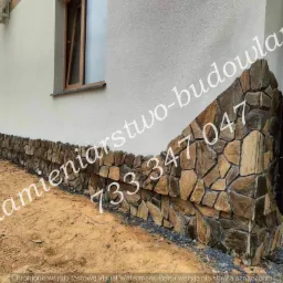 Kamienna okładzina elewacyjna na cokole budynku, wykonana z nieregularnych, brązowo-szarych kamieni, z widoczną rynną spustową i fragmentem przyczepy kempingowej w tle.