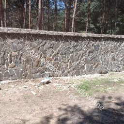 Kamieniarstwo- budowlane i inne usługi - Kamienne ogrodzenie z nieregularnych, szarych kamieni z jasną fugą, zwieńczone płaskimi kamieniami. W tle las iglasty, ziemia z trawą u podstawy.