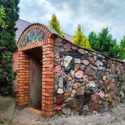 Kamieniarstwo- budowlane i inne usługi - Kamienna ściana z ceglanym wejściem, zwieńczona łukiem z kamienia i cegły. Tło stanowią drzewa i krzewy, a całość prezentuje się jako element architektury ogrodowej.