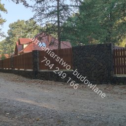 Kamieniarstwo -budowlane - Długi, drewniany płot z kamiennymi słupkami, otaczający posesję z domem o czerwonym dachu, widziany z perspektywy drogi gruntowej.
