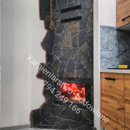 Kamieniarstwo- budowlane i inne usługi - Kamienny kominek z paleniskiem, obudowany nieregularnym kamieniem z dekoracyjnym sznurem. Widoczne płomienie i fragment zabudowy kuchennej.