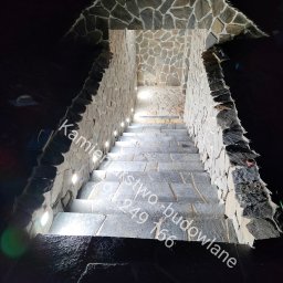 Kamieniarstwo- budowlane i inne usługi - Kamienne schody zewnętrzne z oświetleniem punktowym w ścianach, prowadzące w górę, wykończone nieregularnym kamieniem. Ujęcie z dołu, ciemne tło.