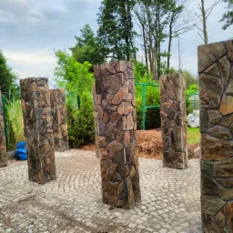 Kamienne słupki ogrodzeniowe z nieregularnych płyt, ułożone na kostce brukowej, widok na zieleń ogrodu w tle.