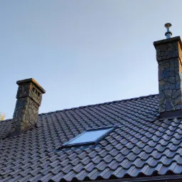 Dwa kominy wykończone kamieniem na brązowym dachu z dachówką ceramiczną i oknem dachowym, widok z dołu na tle jasnego nieba.