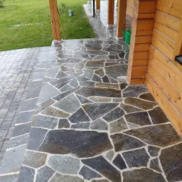 Kamienny taras przy drewnianym domu, z nieregularnych płyt w odcieniach szarości i brązu, widoczny fragment trawnika i konewka.