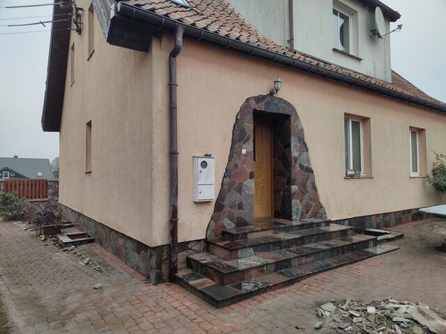 Wejście do domu z kamiennymi schodami i obramowaniem drzwi w kształcie łuku, wykonane z nieregularnych kamieni w odcieniach szarości i czerwieni, z widocznymi pozostałościami po pracach budowlanych...