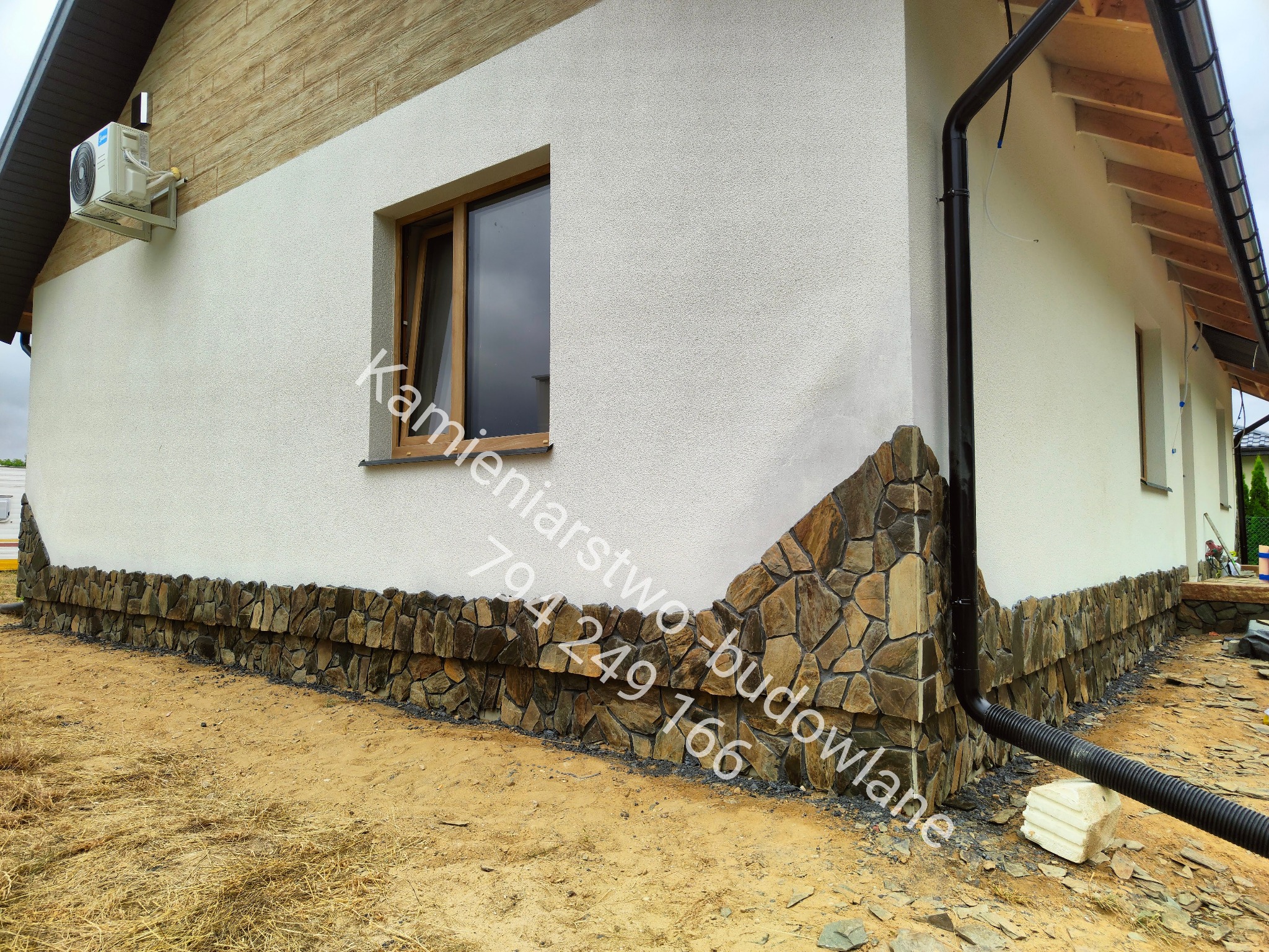 Wykończenie elewacji domu kamieniem naturalnym w odcieniach brązu, widoczne okno i rynna. Częściowo wykończona elewacja, surowy teren wokół budynku.