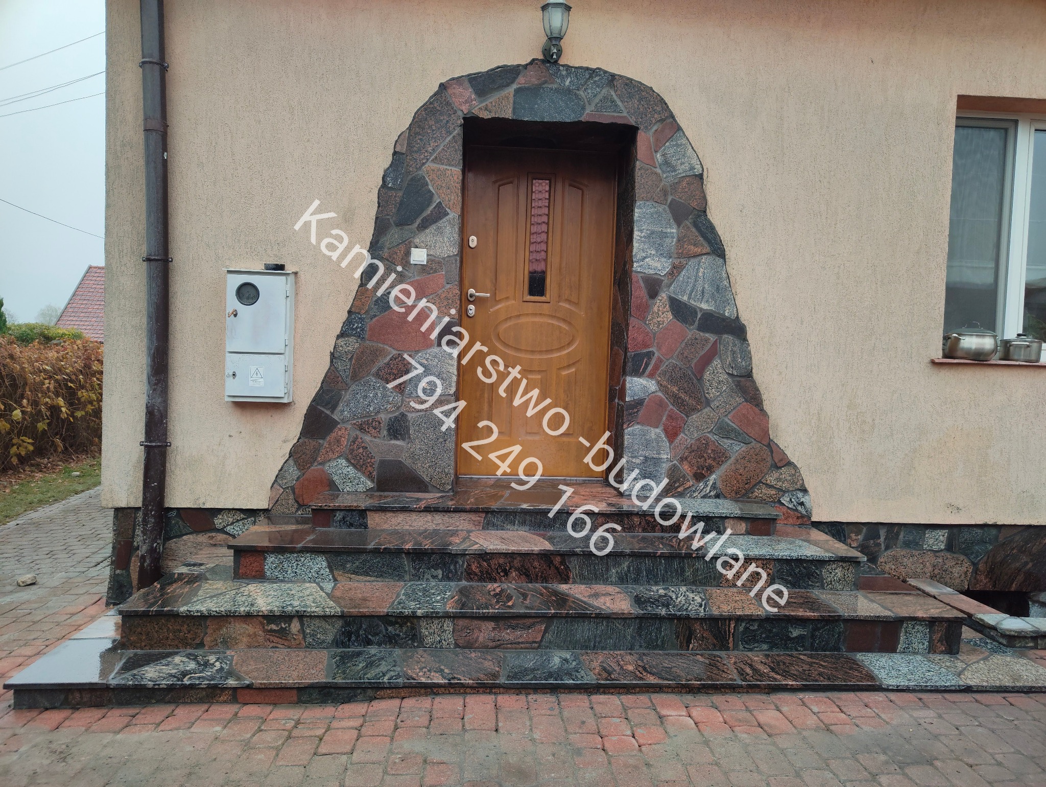 Kamienne schody wejściowe z obudową drzwi z nieregularnych kamieni w odcieniach szarości i czerwieni, prowadzące do drewnianych drzwi wejściowych.