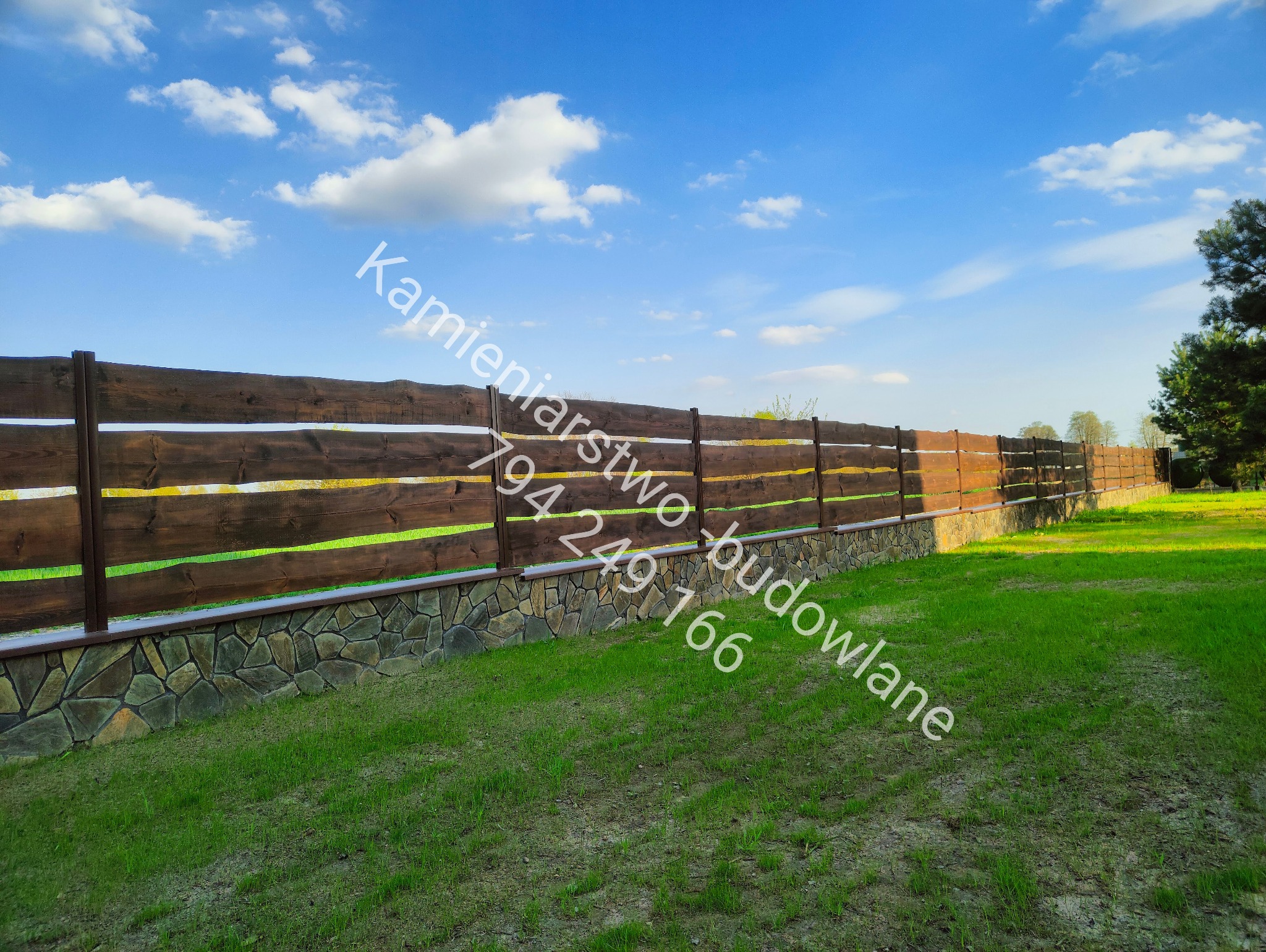Drewniane ogrodzenie poziome z podmurówką z kamienia naturalnego na tle błękitnego nieba z chmurami i zielonego trawnika. Widok z poziomu gruntu.