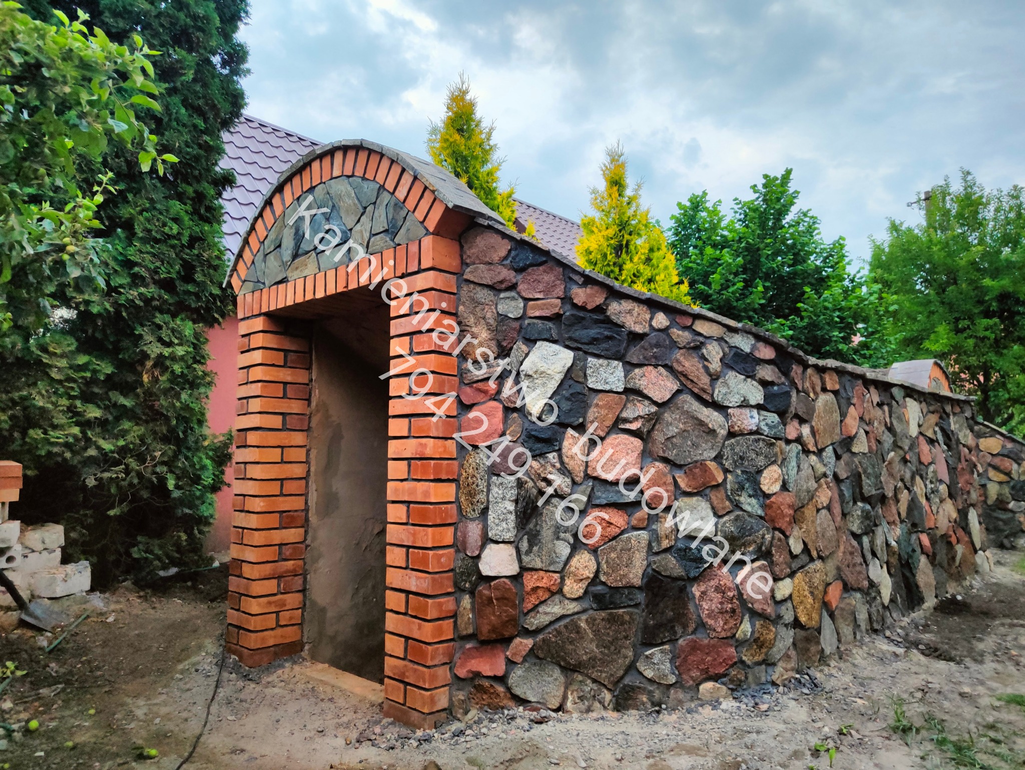 Kamienna ściana z ceglanym wejściem, zwieńczona łukiem z kamienia i cegły. Tło stanowią drzewa i krzewy, a całość prezentuje się jako element architektury ogrodowej.