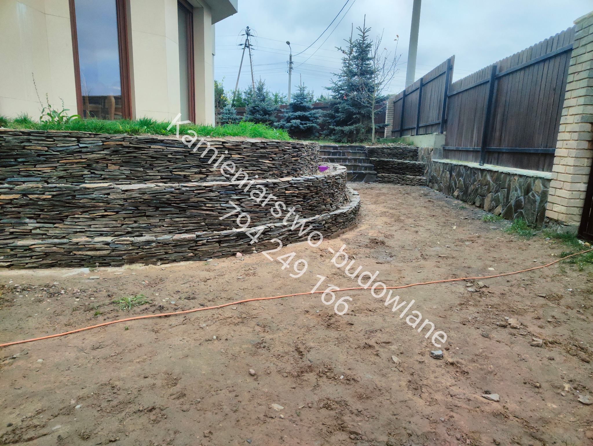 Kamienne tarasy kaskadowe z roślinnością i schodami, otoczone zielenią i ogrodzeniem. Widok na dom z brązowymi oknami. Pochmurne niebo.