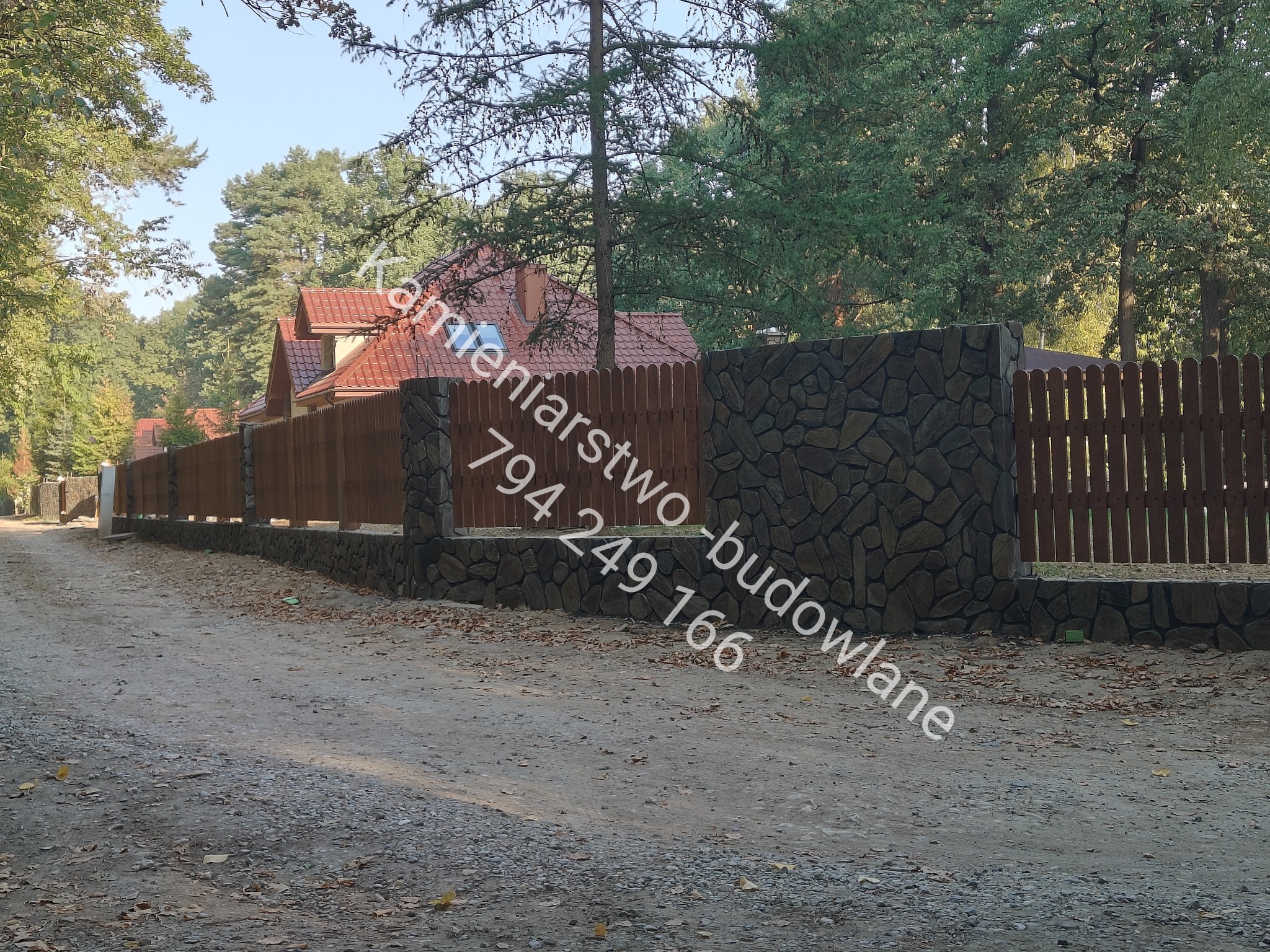 Długi, drewniany płot z kamiennymi słupkami, otaczający posesję z domem o czerwonym dachu, widziany z perspektywy drogi gruntowej.