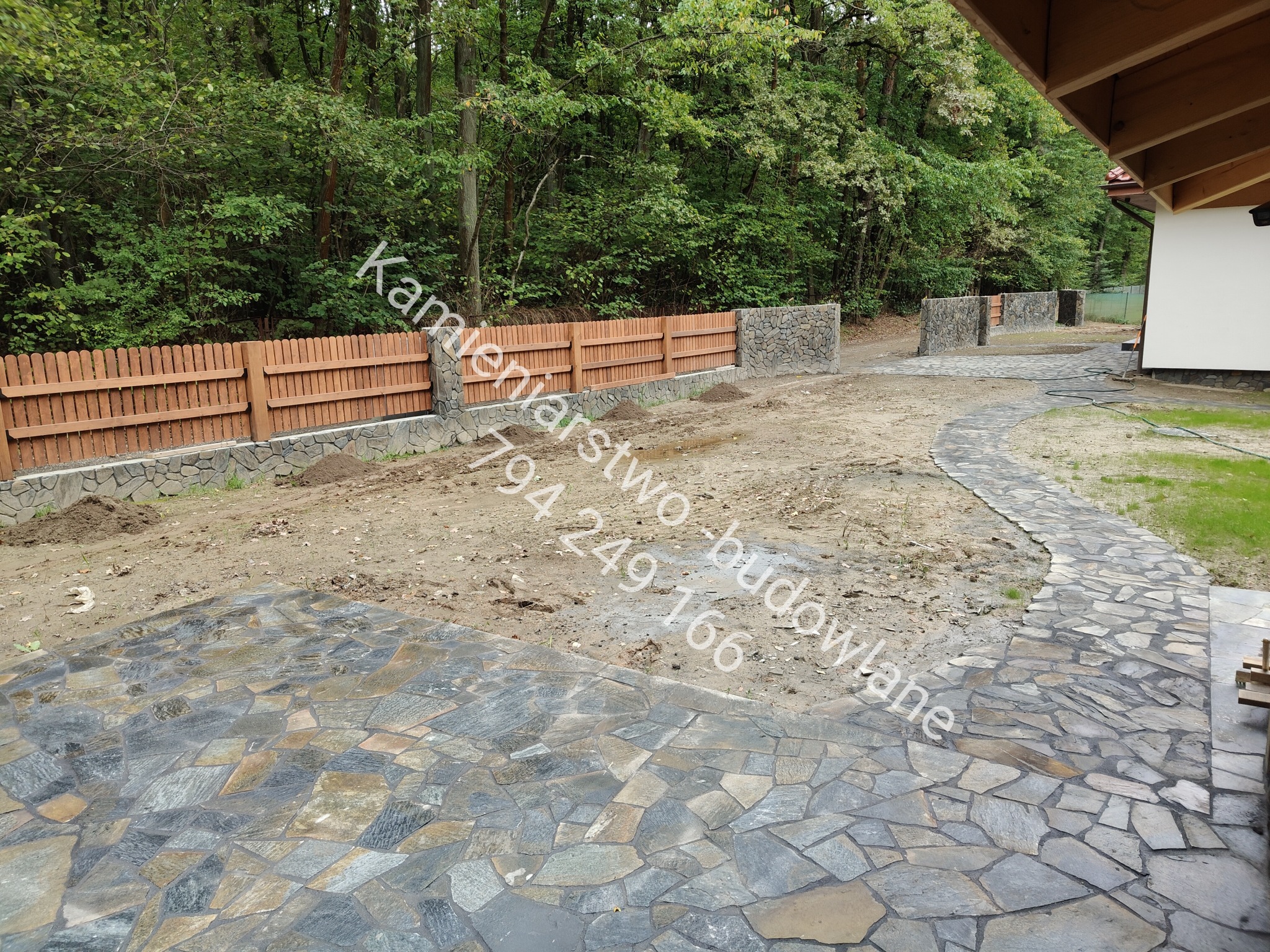 Wykończony taras z kamienia naturalnego i drewniano-kamienne ogrodzenie na tle zieleni. Ścieżka z kamienia prowadzi wzdłuż domu. Widoczne elementy konstrukcyjne dachu.