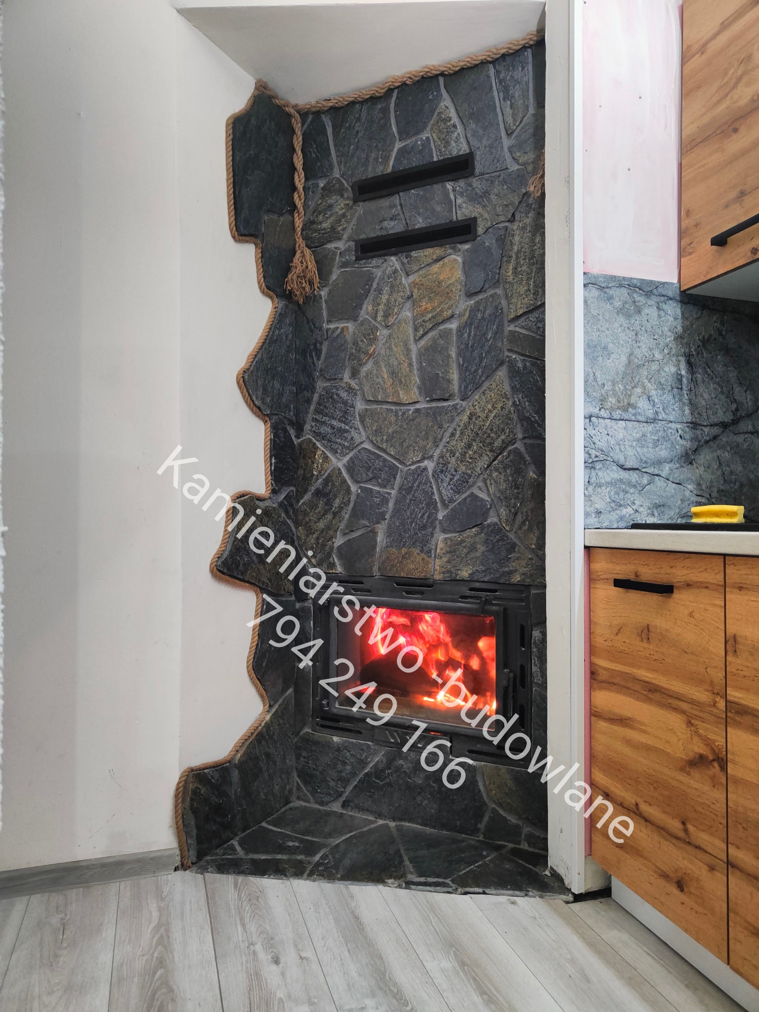 Kamienny kominek z paleniskiem, obudowany nieregularnym kamieniem z dekoracyjnym sznurem. Widoczne płomienie i fragment zabudowy kuchennej.
