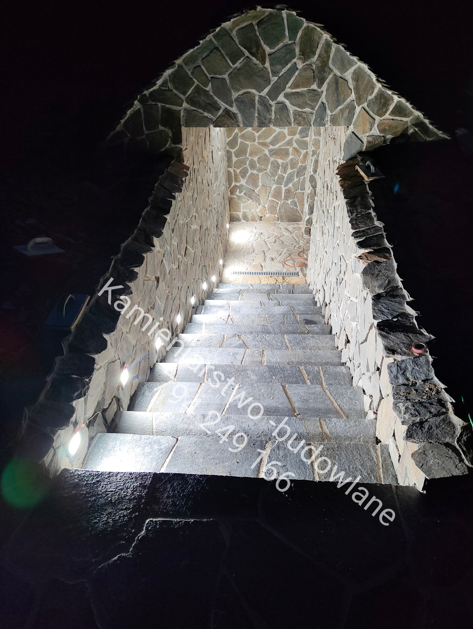 Kamienne schody zewnętrzne z oświetleniem punktowym w ścianach, prowadzące w górę, wykończone nieregularnym kamieniem. Ujęcie z dołu, ciemne tło.