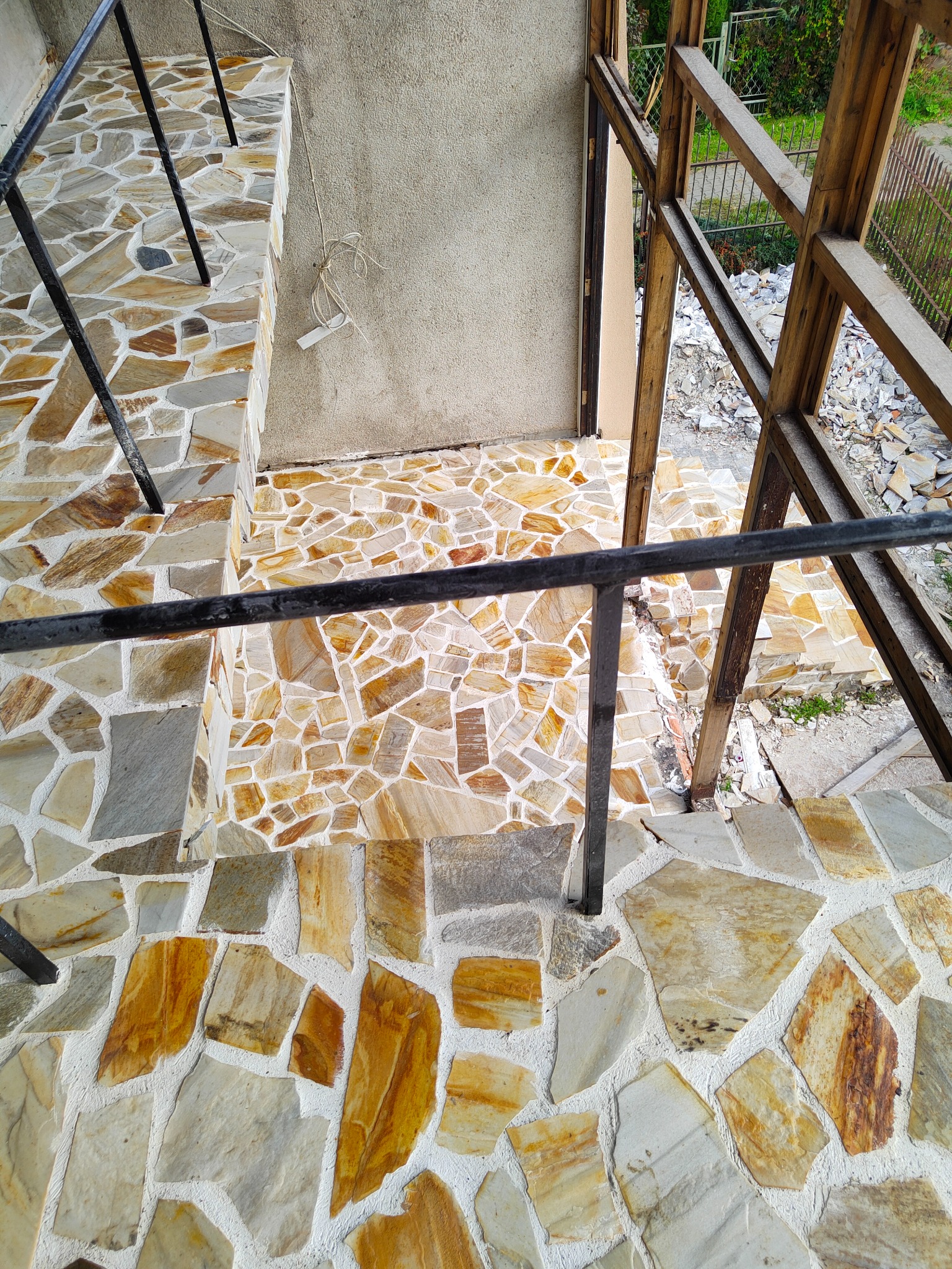 Schody i taras wyłożone nieregularnymi płytami kamiennymi w odcieniach beżu i szarości, czarna metalowa balustrada, widoczne pozostałości materiału budowlanego i drewniana konstrukcja szkieletowa...