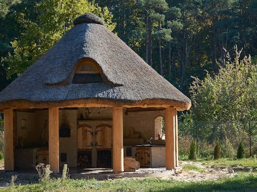 Zadaszona letnia kuchnia z cegły z piecem chlebowym i grillem, kryta strzechą z małym okrągłym oknem, otoczona zielenią.