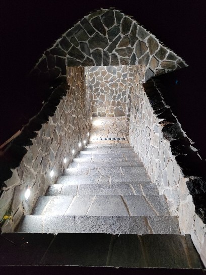 Kamienne schody zewnętrzne z oświetleniem punktowym wbudowanym w ściany boczne, prowadzące w górę pod kamiennym zadaszeniem, nocne ujęcie.