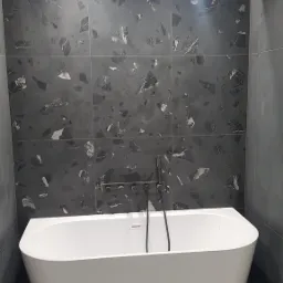 Nowoczesna łazienka z białą wanną wolnostojącą na tle ściany wyłożonej ciemnoszarymi płytkami imitującymi lastryko, oświetlona punktowymi lampami.