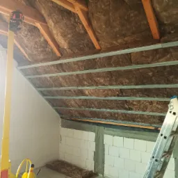Poddasze w trakcie ocieplania wełną mineralną, widoczne metalowe profile pod płyty gipsowo-kartonowe, poziomica laserowa na statywie oraz drabina aluminiowa.