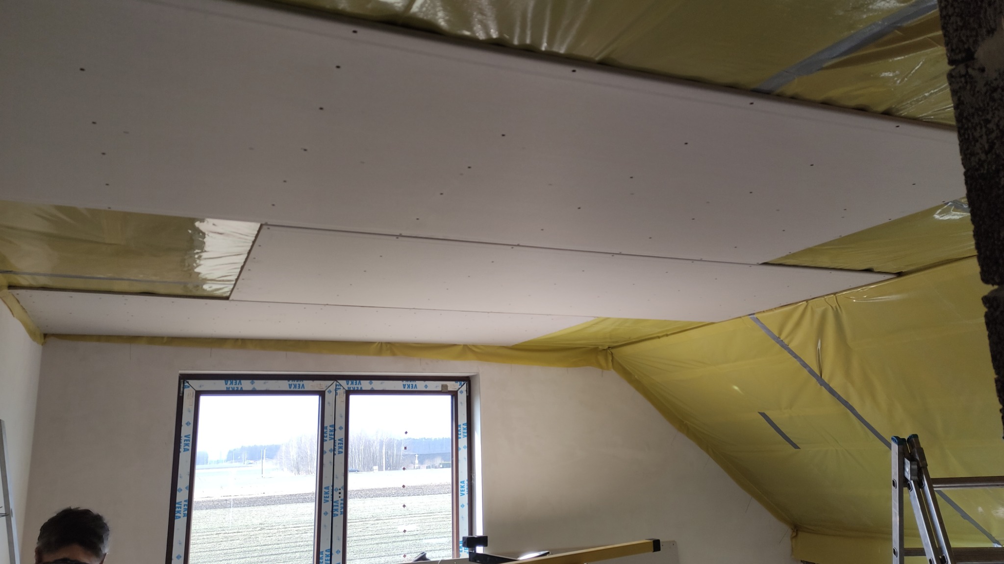 Montaż sufitu podwieszanego z płyt karton gips, widoczne płyty przymocowane do stelaża, okno z widokiem na pola, folia paroizolacyjna w żółtym kolorze oraz fragment drabiny.