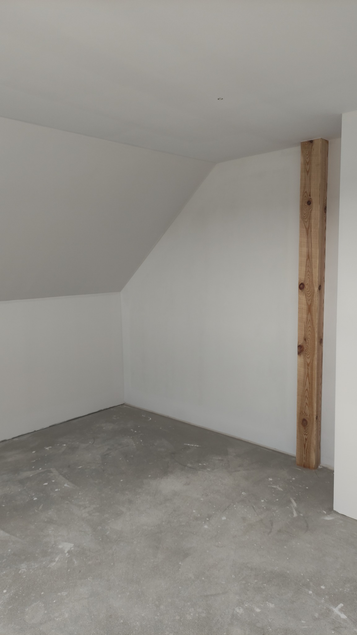 Wnętrze pokoju na poddaszu z surowym betonem na podłodze, świeżo pomalowanymi na biało ścianami i widoczną drewnianą belką konstrukcyjną o wyraźnym usłojeniu.