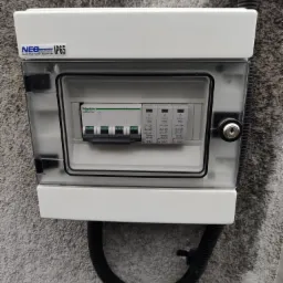 Zamknięta skrzynka elektryczna z przezroczystym wiekiem, zawierająca wyłączniki nadprądowe i ograniczniki przepięć, zamontowana na szarej, chropowatej ścianie zewnętrznej z widoczną rurą karbowaną...