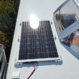 Panel słoneczny z okablowaniem zamontowany na dachu pojazdu, widoczny otwarty świetlik dachowy i drabina w tle.