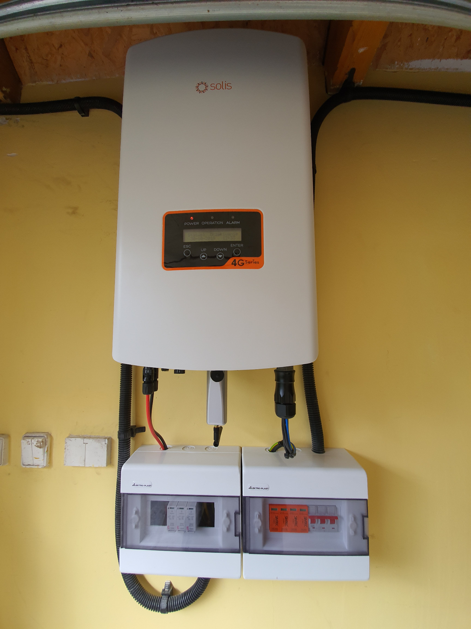 Biały falownik Solis 4G Series zamontowany na żółtej ścianie, z widocznymi przewodami i dwoma skrzynkami bezpiecznikowymi Elektro-Plast poniżej, całość instalacji wewnątrz budynku.