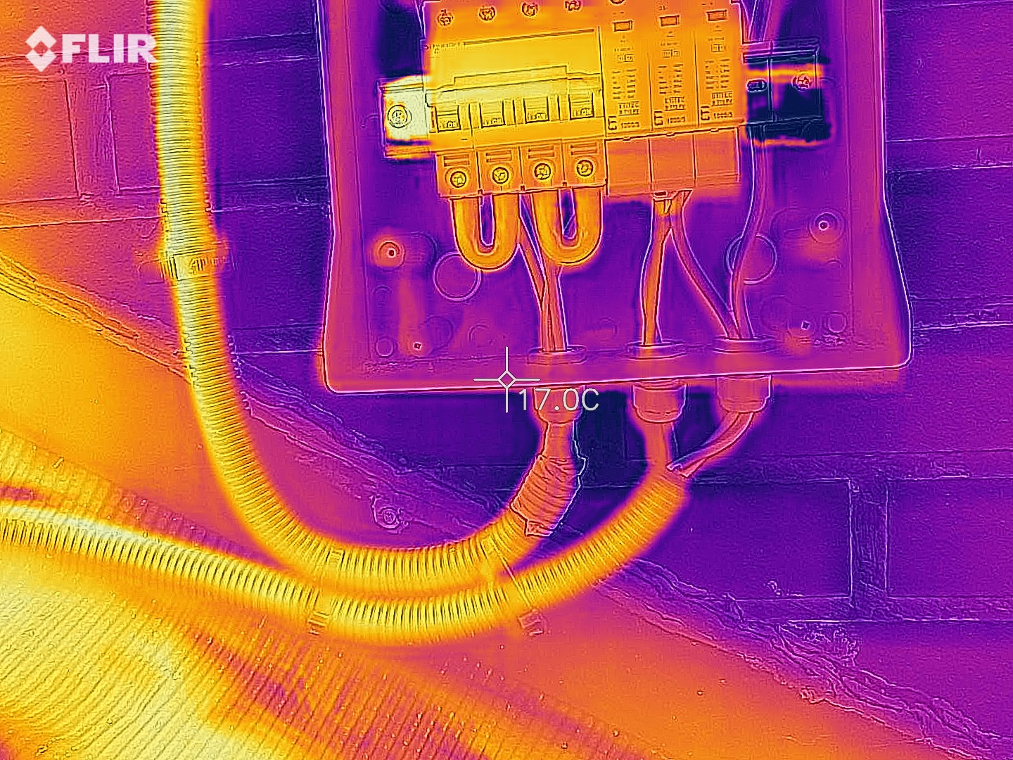 Termowizyjne zdjęcie instalacji elektrycznej w skrzynce z bezpiecznikami, widoczne kable i przewody o różnej temperaturze, pomiar temperatury wskazuje 17 stopni Celsjusza, tło z cegieł.