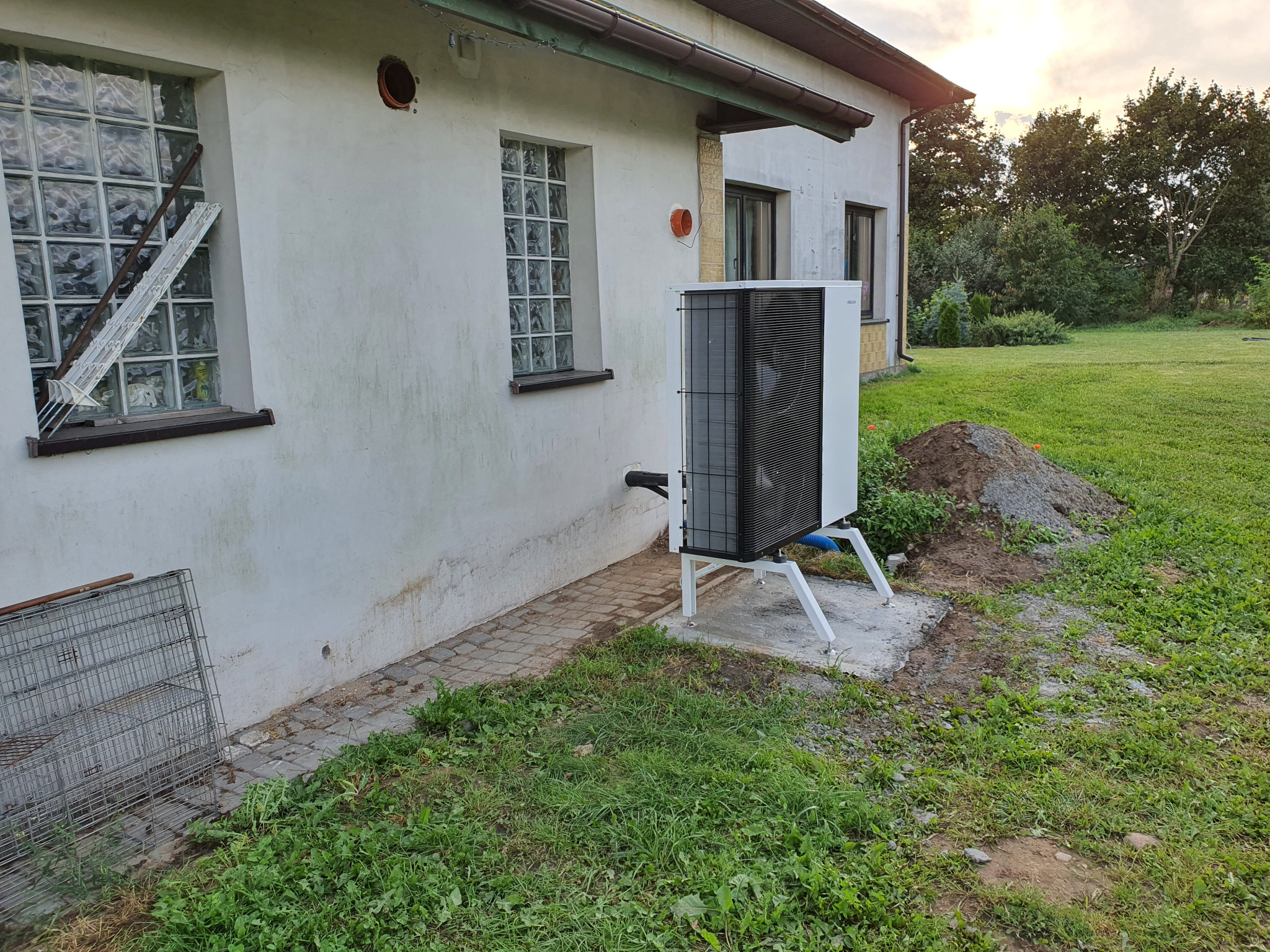 Biała pompa ciepła powietrze-woda zamontowana na zewnątrz budynku z oknami z luksferów i nierównym trawnikiem, widoczna mała górka ziemi.