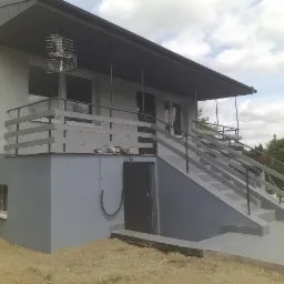 Dwukondygnacyjny dom z szarą elewacją, schodami wejściowymi z metalowymi poręczami i anteną telewizyjną na dachu.