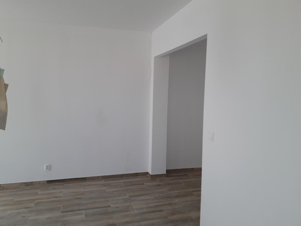 Wnętrze świeżo wykończonego pokoju z gładkimi, białymi ścianami, drewnopodobną podłogą i widocznym wejściem do kolejnego pomieszczenia.