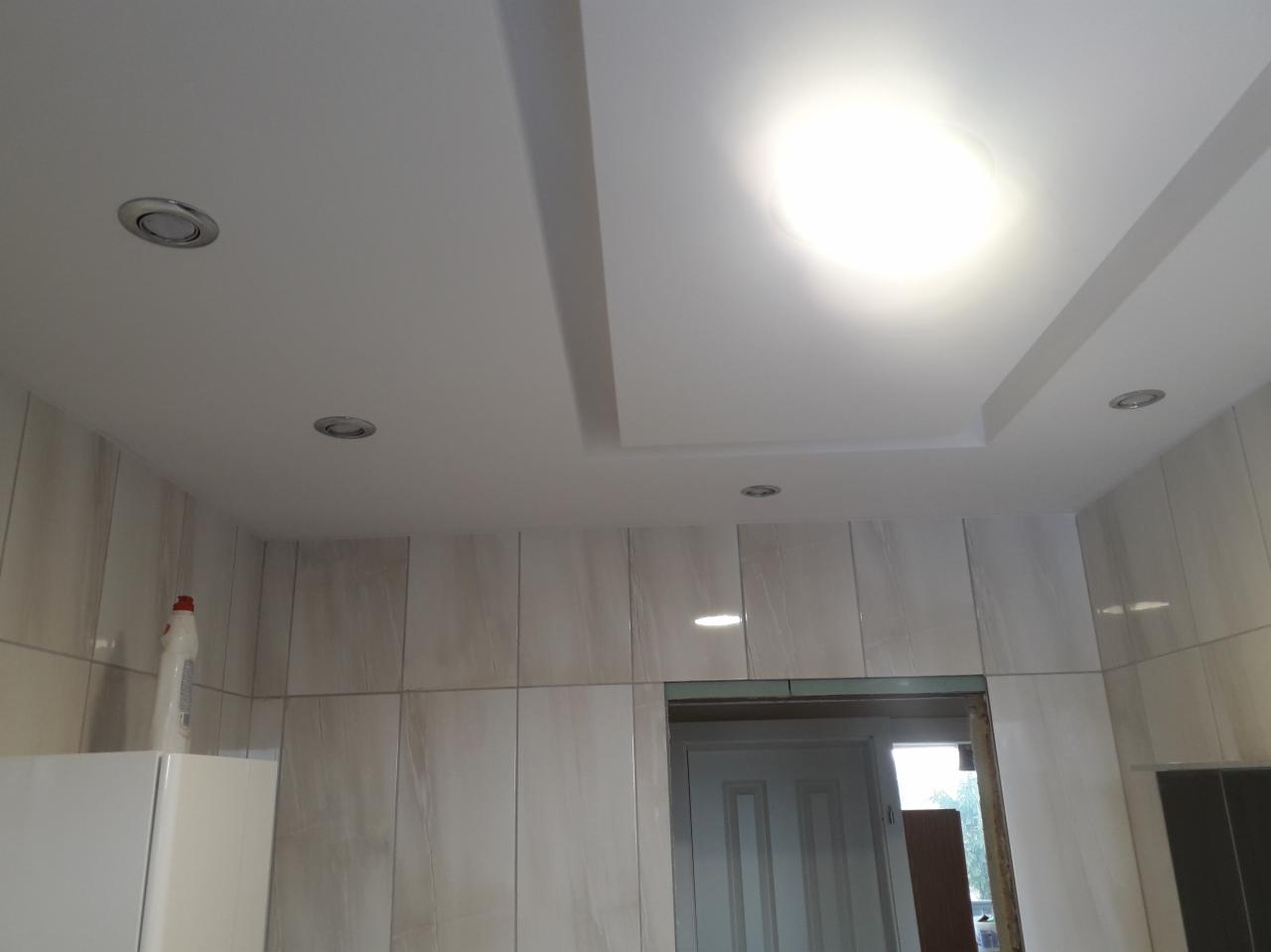 Wykończony sufit podwieszany z oświetleniem punktowym w pomieszczeniu z okładziną ceramiczną imitującą marmur, widoczny fragment białej lodówki i butelka z płynem.